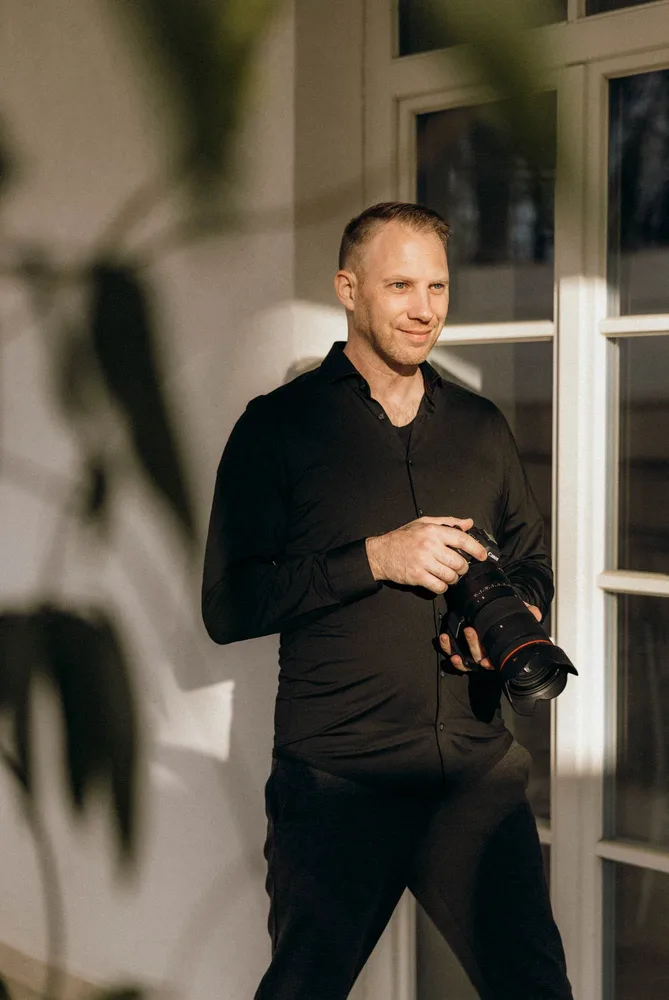 Andy, Fotograf und Videograf in schwarzem Hemd und Hose hält eine Kamera mit großem Objektiv. Hochzeitsfotograf von MOKATI