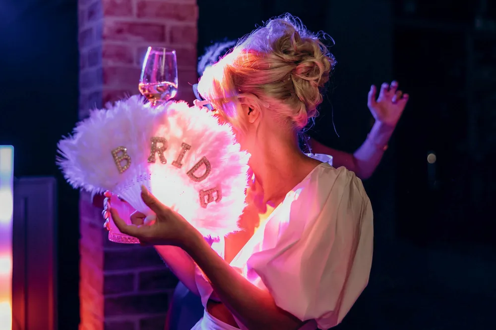 Blonde Braut in weißem Kleid hält einen leuchtenden Federfächer bei der Hochzeit in der Alten Gärtnerei München