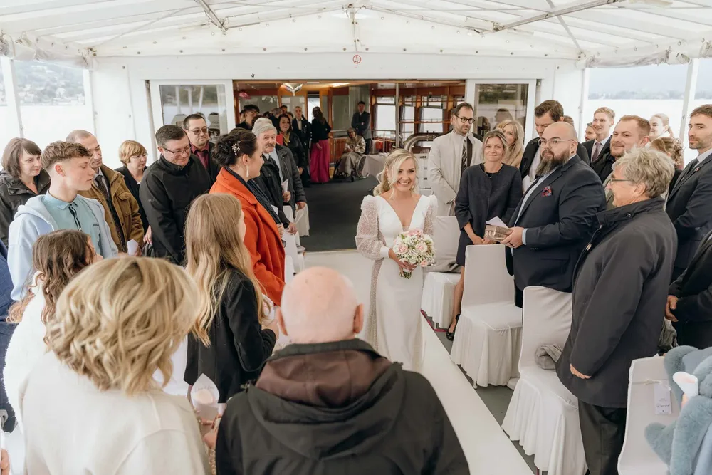 Braut mit Brautstrauß geht auf einem weißen Teppich durch Hochzeitsgesellschaft auf dem Schiff am Tegernsee