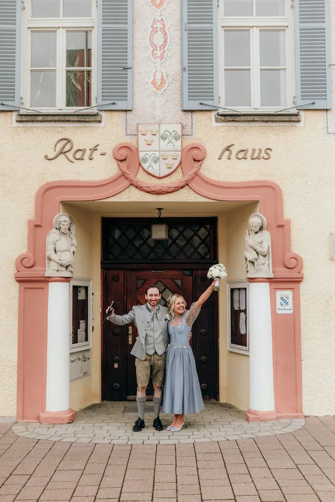 Brautpaar steht am Flussufer vor einem gelben Gebäude beim Rathaus Tegernsee