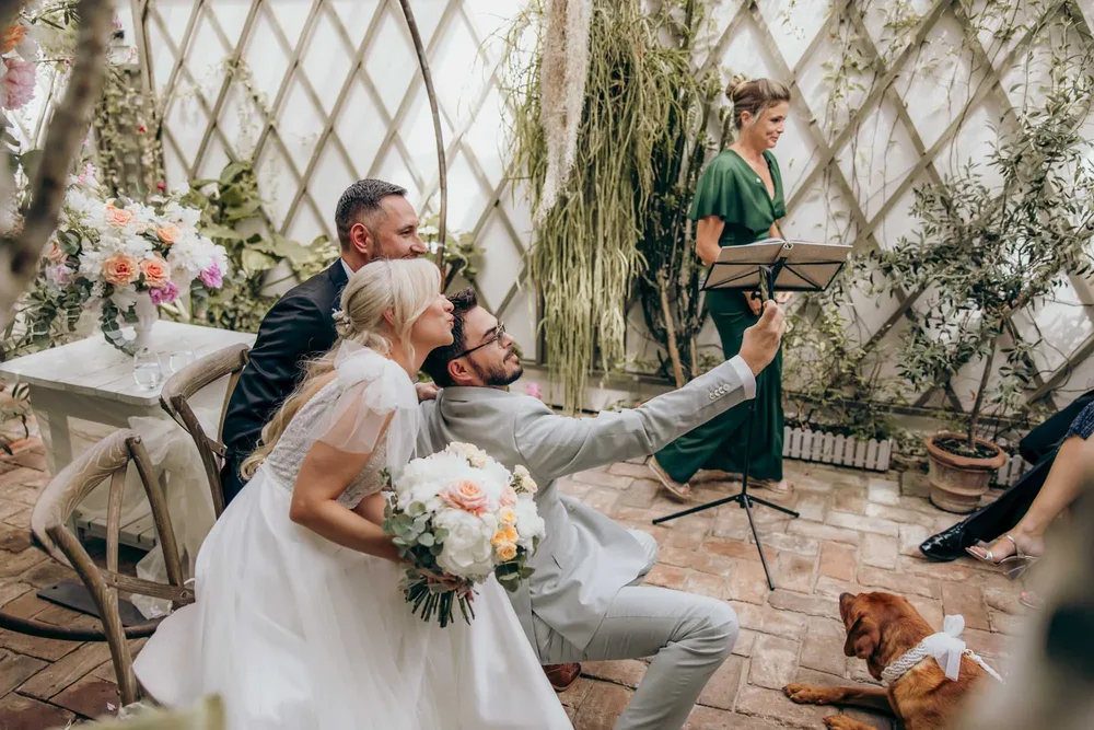Brautpaar und Gast machen ein Selfie, ein Hund liegt auf dem Boden. Hochzeitsfotograf München MOKATI