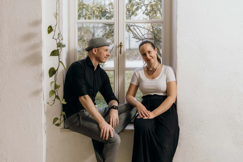 Claudia und Andy sitzen an einem Fenster, umgeben von Pflanzen. Das sind die Hochzeitsfotografen-Duo von MOKATI