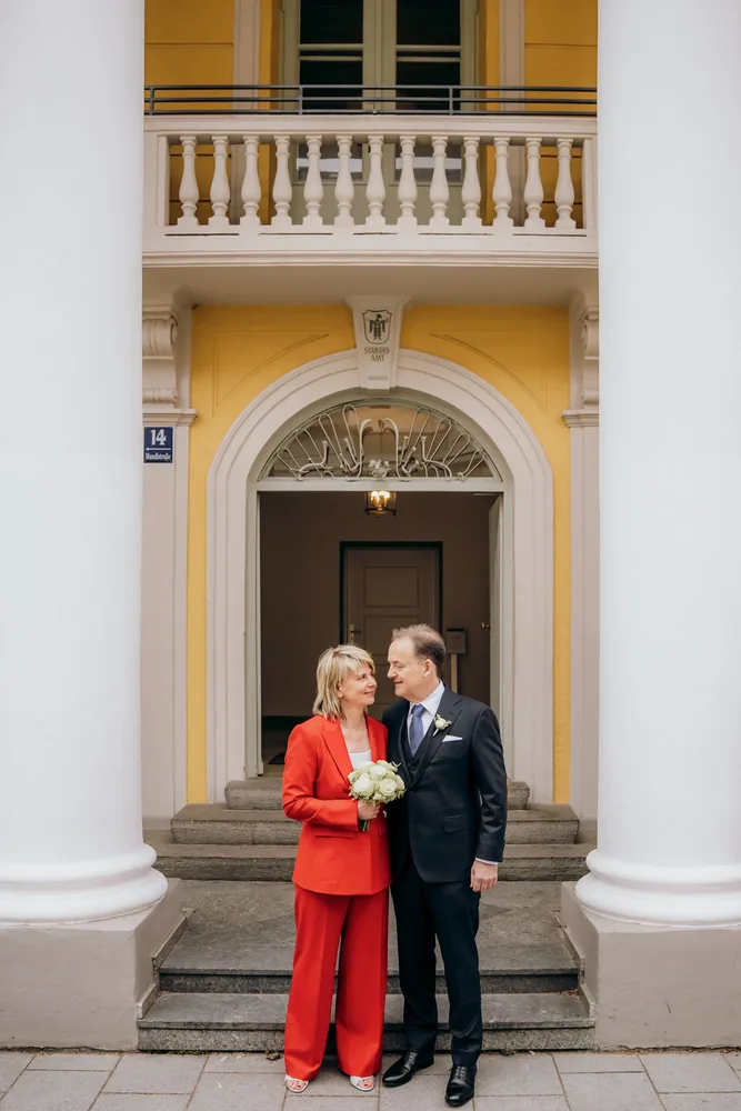 Lächelndes Brautpaar, sie in rotem Hosenanzug mit Blumen, fotografiert von MOKATI