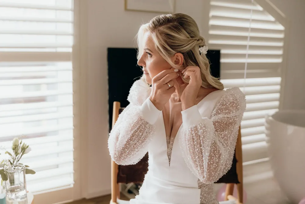 Eine blonde Frau sitzt vor einem Spiegel, während ein weißes Hochzeitskleid an einem Holzbalken hängt. Hochzeitsfotograf München MOKATI