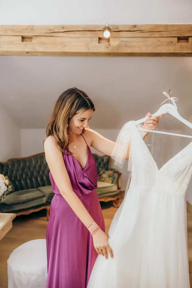Lächelnde Frau in violettem Kleid betrachtet ein Brautkleid beim Getting Ready