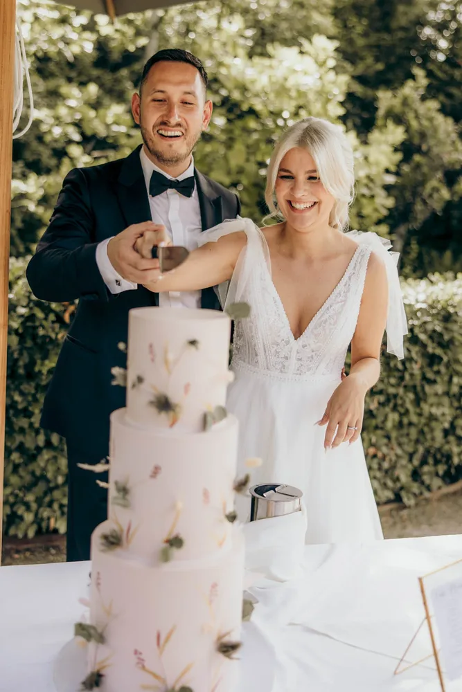 Lächelndes Brautpaar schneidet gemeinsam eine dreistöckige Hochzeitstorte in der Alten Gärtnerei München an
